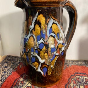 May include: A ceramic jug with a dark brown base and a decorative pattern of gold, blue, and white. The jug has a handle and a spout for pouring. The design is abstract and artistic, suitable for serving beverages.