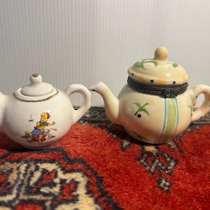 May include: Two miniature ceramic teapots. The left teapot is white with a gold rim and a colourful illustration of a person cooking. The right teapot is cream-coloured with green stripes, blue dots, green leaf accents, and a dark metal hinge.