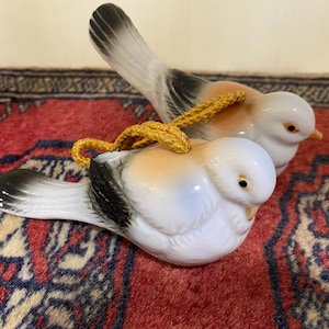 May include: Two ceramic bird ornaments. The birds have white bodies, black and grey wings, and orange and tan details. A gold-coloured cord is attached to the top of each bird. The birds are on a red and blue patterned rug.