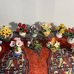 May include: A selection of miniature ceramic flower arrangements, including roses, lilies, and daisies, in various vases and baskets. The flowers are in shades of red, yellow, purple, and white, set on a patterned rug.