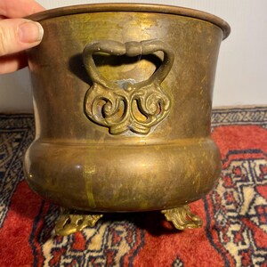 May include: A round brass planter with a weathered patina, featuring two ornate, scroll-shaped side handles and three decorative feet. The metal surface shows signs of oxidation and discolouration, with a wide opening and a curved body shape.