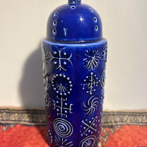 May include: A cylindrical cobalt blue ceramic jar with a matching domed lid. The exterior surface features a repeating pattern of white embossed geometric symbols, including sunbursts, spirals, and cross-like motifs, with a glossy finish.