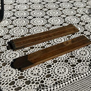 May include: Two vintage folding wooden rulers with a dark brown finish and metal hinges. The rulers are rectangular, showing a smooth wood grain texture and visible joint mechanisms at the ends. These measuring tools are positioned diagonally.