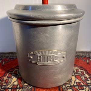 May include: A cylindrical brushed aluminium storage canister with a fitted lid and a small red knob handle. A rectangular metal plaque is attached to the front, featuring the word RICE embossed in capital letters with a decorative border design.