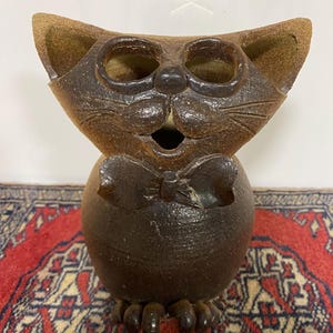 May include: A brown ceramic cat figurine with a bow tie. The cat has large eyes and a wide open mouth. The figurine is sitting on a red and cream rug.