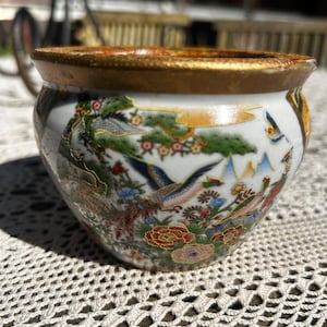 May include: A small, round porcelain bowl featuring a gold-painted rim and a detailed, colourful scene of birds, pine trees, and blooming flowers. The white background highlights the intricate illustrations of pheasants and floral patterns.