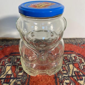 May include: A clear glass bear-shaped jar with a blue lid, featuring the words "Easy Cheese". The jar is displayed on a red patterned rug. The jar is a vintage item.
