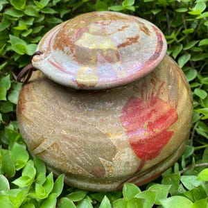 May include: A ceramic jar with a lid, decorated with a floral design in shades of red, gold, and green. The jar has a rustic, handmade look.