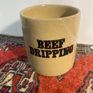 May include: A beige ceramic container with the words "BEEF DRIPPING" in bold, dark brown letters. The container is cylindrical with a slightly flared rim. It sits on a red and blue patterned rug.