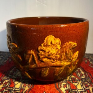 May include: A round, brown ceramic bowl featuring a raised, textured relief design of flowers and foliage wrapping around the exterior. The glaze has a glossy finish with visible fine crackling throughout the surface of the dark brown pottery.