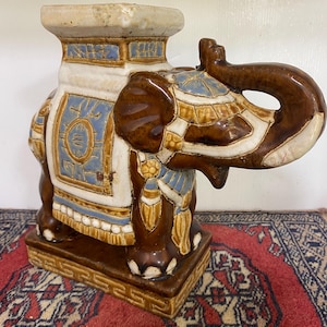 May include: A ceramic elephant figurine with a square base and intricate details. The elephant is brown with blue and gold accents. The figurine is a decorative piece for a home or garden.