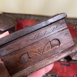 May include: A small, dark brown wooden box with carved floral and leaf designs. The box has a rectangular shape and a hinged lid. The wood appears aged, with visible grain and a slightly rough texture. The box is held in a hand.