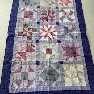 May include: A rectangular quilt featuring a grid of fifteen square blocks in shades of purple, pink, and white. Each block displays a unique geometric pattern. The quilt is framed by a light purple border and a wide, dark purple outer border.