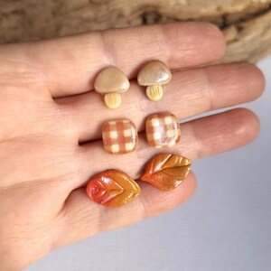 May include: A collection of handmade earrings in the shape of mushrooms, plaid squares, and autumn leaves. The mushroom earrings are beige with light brown stems. The plaid earrings are red and white. The leaf earrings are orange and brown.
