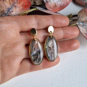 Puede incluir: Un par de pendientes ovalados dorados con un diseño veteado e iridiscente. Los pendientes tienen un marco dorado y un pequeño perno redondo dorado en la parte superior. El diseño veteado presenta tonos de gris, marrón y plata con acentos de purpurina.