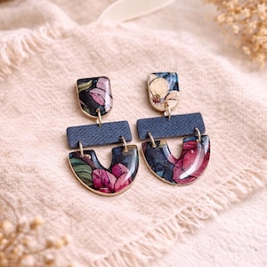 Könnte beinhalten: Blumen-Ohrhänger mit einem marineblauen, jeansartigen Akzent. Die Ohrringe haben einen rechteckigen oberen Teil und einen U-förmigen unteren Teil mit goldfarbenem Rand. Das florale Design umfasst rosa, rote und grüne Farbtöne auf dunklem Hintergrund.