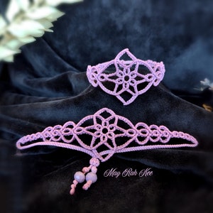 以下が含まれることがあります： 花柄の繊細なピンクのクロシェ編みブレスレット2点セット。ブレスレットは黒い布地に置かれています。大きなブレスレットにはビーズのタッセルが付いています。