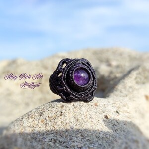 Può includere: Un anello in macramè nero con una pietra preziosa di ametista viola. L'anello è su una superficie sabbiosa. Il testo "May Rah Kee Amethyst" è visibile nell'immagine.