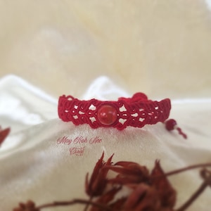 May include: Red braided bracelet with a red bead in the center. The bracelet is on a white fabric background.