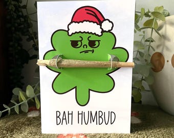 May include: A white greeting card featuring a green, grumpy-faced cartoon character wearing a red and white Santa hat. The character holds a rolled paper cylinder across its center. The text "BAH HUMBUD" is printed in black at the bottom.