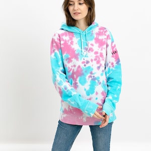 Frontside of a baby blue baby pink tie dye hoodie on a model standing in front of white background. Tie dye sweatshirt is with colorful transgender pattern design. Jumper is with long sleeves hood small listee logo on the sleeve.