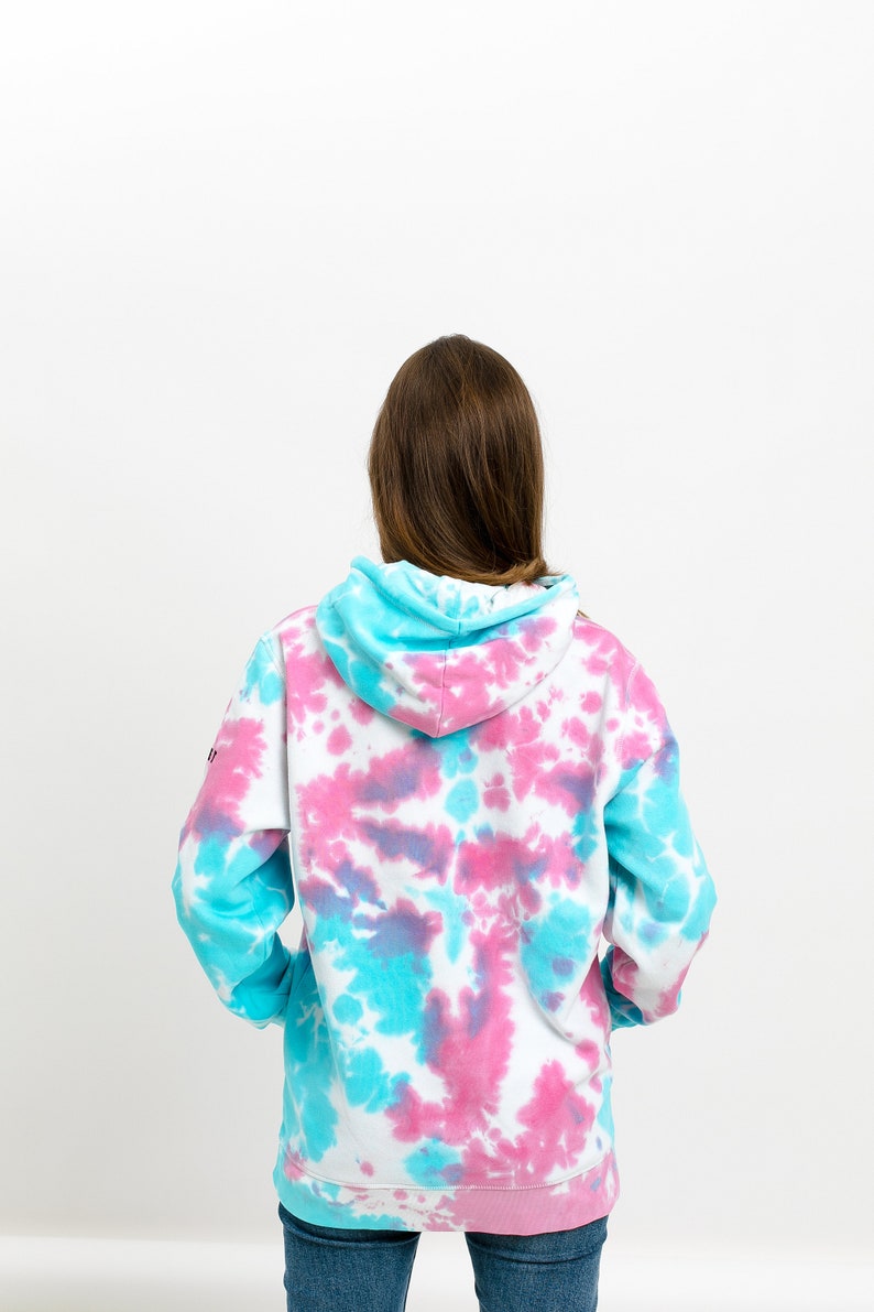 Backside of a baby blue baby pink tie dye hoodie on a model standing in front of white background. Tie dye sweatshirt is with colorful transgender pattern design. Jumper is with long sleeves hood small listee logo on the sleeve.