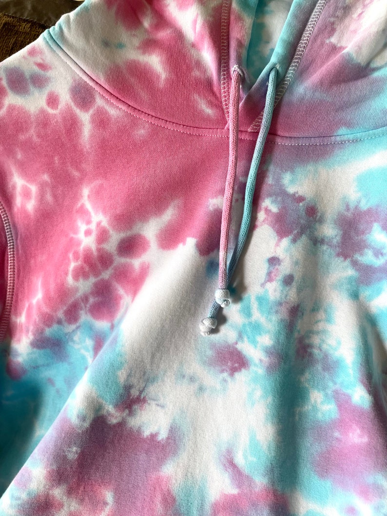 Details of a tie dye baby blue and baby pink hoodie. Close up image of hood straps and hoodie stiches. Hoodie made from soft organic cotton material.