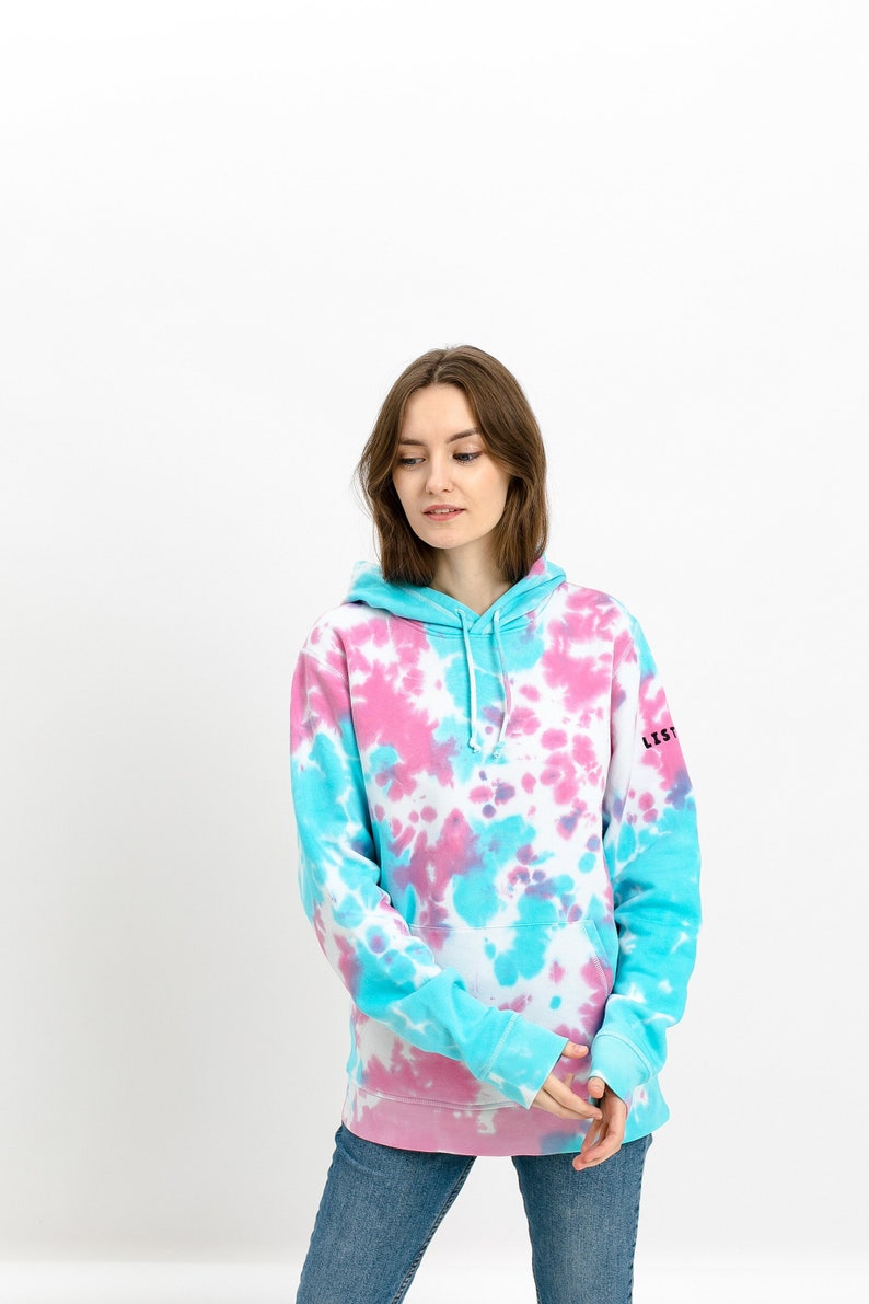 Frontside of a baby blue baby pink tie dye hoodie on a model standing in front of white background. Tie dye sweatshirt is with colorful transgender pattern design. Jumper is with long sleeves hood small listee logo on the sleeve.