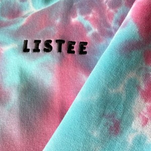 Details of a tie dye baby blue and baby pink hoodie. Close up image of embroidered black logo on the sleeve. Hoodie made from soft organic cotton material.