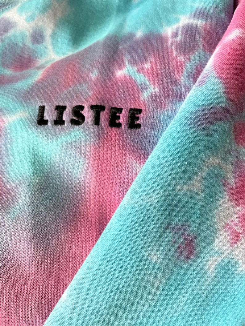 Details of a tie dye baby blue and baby pink hoodie. Close up image of embroidered black logo on the sleeve. Hoodie made from soft organic cotton material.