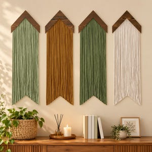 May include: Four wall hangings featuring dark brown wooden chevron tops with long, straight hanging yarn strands in sage green, mustard yellow, sage green, and cream. Each piece has a pointed bottom edge, creating a uniform arrow-like shape.