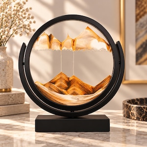 May include: A circular black frame holds a glass display containing layers of golden and white sand that form mountain-like shapes. The frame is mounted on a rectangular black base, allowing the sand to shift and create new patterns within the glass.