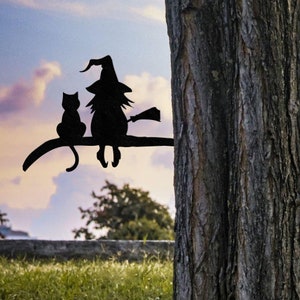 Könnte beinhalten: Schwarze Metallsilhouette einer Hexe und einer Katze, die auf einem Ast sitzen. Die Silhouette ist an einem Baumstamm befestigt.