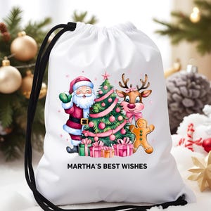 Puede incluir: Bolsa blanca con cordón con un diseño navideño. La ilustración incluye a Papá Noel, un reno, un árbol de Navidad y un hombre de jengibre. El texto "MARTHA'S BEST WISHES" está impreso en la bolsa. Cierre de cordón negro.