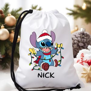 Può includere: Borsa bianca con coulisse con un personaggio dei cartoni animati con un cappello da Babbo Natale e luci natalizie. Il personaggio è blu con grandi orecchie e una sciarpa rossa. Il nome "NICK" è stampato in nero.