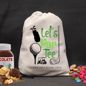 May include: A cream drawstring bag with a golf theme. The bag displays a golf ball on a tee, a golf cart, and the words "Let's Par Tee" in green. The text "LUCAS'S LAST ROUND 2024" is also printed on the bag. Chocolate and sweets are in the foreground.