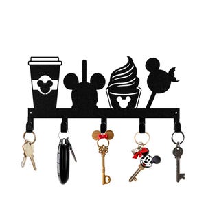 Può includere: Portachiavi in metallo nero con cinque ganci. Il portachiavi è a tema Topolino con silhouette di una tazza di caffè, orecchie di Topolino, un cono gelato di Topolino e un lecca-lecca di Topolino. Ci sono cinque chiavi appese ai ganci.