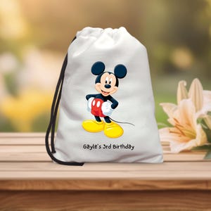 Könnte beinhalten: Weißer Kordelzugbeutel mit einer Mickey Mouse-Illustration. Die Cartoonfigur trägt rote Shorts, gelbe Schuhe und weiße Handschuhe. Der Text "Gayle's 3rd Birthday" ist darunter aufgedruckt. Der Beutel hat einen schwarzen Kordelzug.