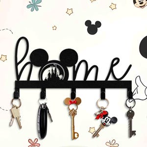 Könnte beinhalten: Schwarzer Schlüsselhalter aus Metall mit dem Wort "home" und Mickey-Maus-Ohren. Der Halter hat fünf Haken und ist mit einer Silhouette von Schloss Aschenputtel verziert.