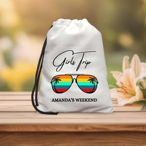 Może przedstawiać: Biały worek ze sznurkiem ze sznurkiem w kolorze czarnym. Torba zawiera napis "Girls Trip" w skrypcie nad parą okularów przeciwsłonecznych z tęczowymi soczewkami i palmami. Pod okularami znajduje się napis "AMANDA'S WEEKEND".