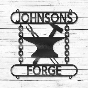 Op de afbeelding: Zwart metalen bord met de tekst "JOHNSONS FORGE" en een silhouet van een aambeeld met een hamer en tang erop. Het aambeeld hangt aan twee kettingen.