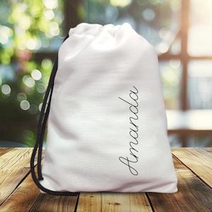 May include: A white drawstring bag with the name "Amanda" in a cursive font. The bag has a black drawstring and is sitting on a wooden surface. The background is blurred, with hints of green and sunlight.