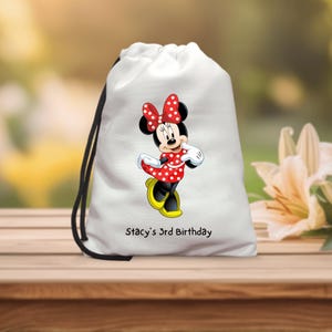 May include: White drawstring bag featuring a Minnie Mouse graphic. Minnie Mouse is wearing a red dress with white polka dots, a matching bow, and yellow shoes. The text "Stacy's 3rd Birthday" is printed on the bag.