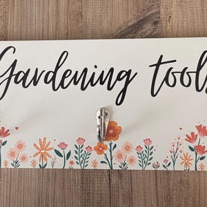 May include: A white wooden sign with the words "Gardening tools" in black script. The sign has three silver hooks and a floral design with orange, pink, and green flowers.