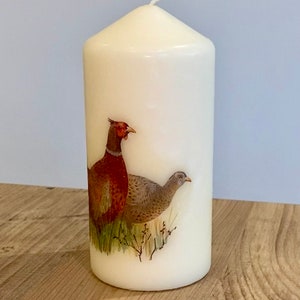 May include: A white pillar candle with a painted image of two pheasants. The pheasants are brown and gray with red accents. The candle is on a wooden surface.
