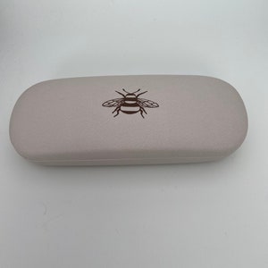 May include: A pale grey glasses case with a textured finish. The case features a brown illustration of a bee on the top. The oval-shaped case is likely made from a robust material.