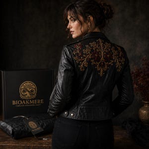 Peut inclure: Une veste en cuir noir ornée d'une broderie florale complexe en fils dorés et rouge foncé sur le haut du dos. La veste présente un col classique, des manches longues et une coupe structurée. Une boîte noire avec le texte doré BIOAKMERE est posée à côté.