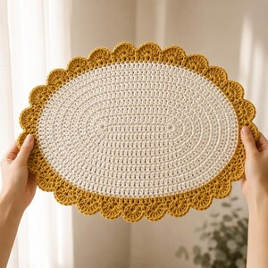 May include: An oval-shaped crochet placemat featuring a cream-colored center with a dense, textured stitch pattern and a wide, scalloped border in a vibrant mustard yellow yarn. The piece has a distinct, decorative edge with rounded loops.