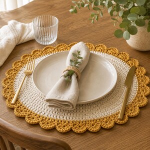 May include: A round crochet placemat featuring a white center and a mustard yellow scalloped border. A beige linen napkin is folded on a white ceramic plate, secured with a woven napkin ring and a small sprig of greenery. A gold fork and knife sit on the placemat.