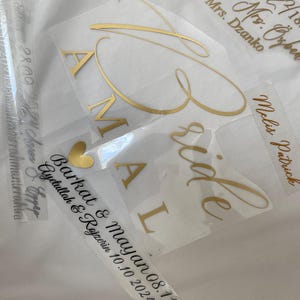 Könnte beinhalten: Mehrere Vinyl-Aufkleber mit goldener und schwarzer Schrift. Die Aufkleber zeigen Namen wie AMAL, Melis Patrick, Barkat & Mayan und Mr. & Mrs. Dzanko sowie verschiedene Daten und ein kleines goldenes Herzsymbol auf einer transparenten Transferfolie.